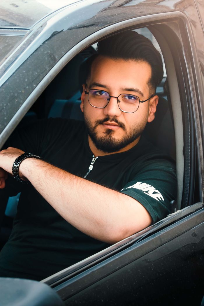 Confident young man wearing glasses sitting in a car, looking directly at the camera.